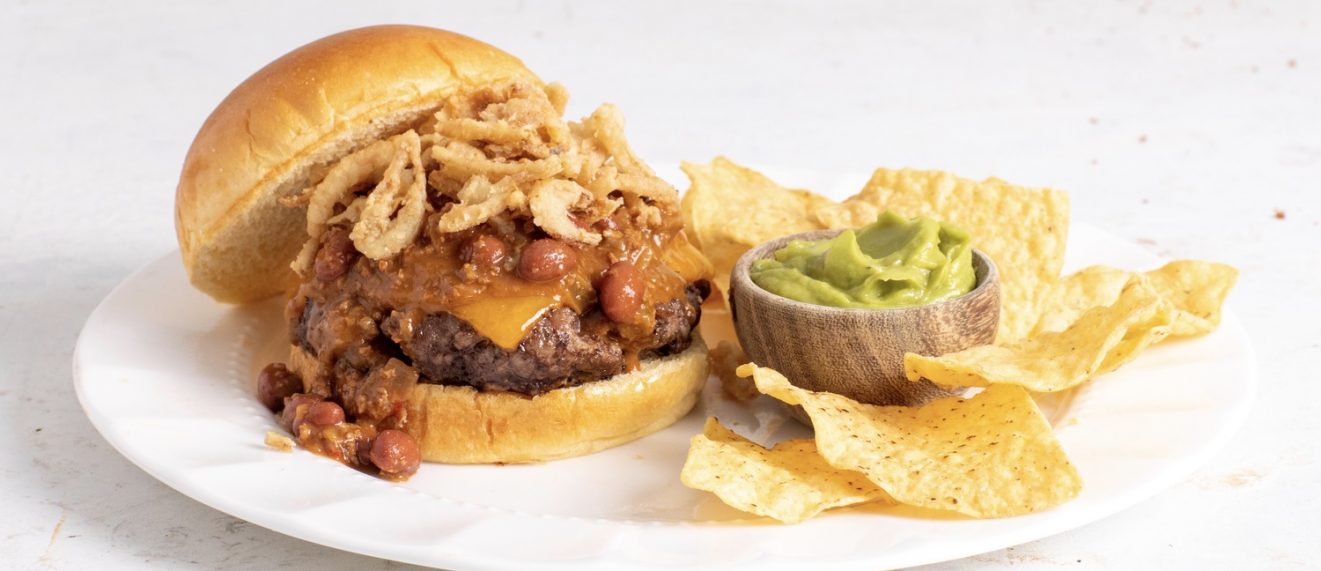 Chili cheeseburger with chips and guacamole sitting on a white plate.