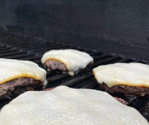 Four single patty burgers topped with cheese cooking on a grated grill.