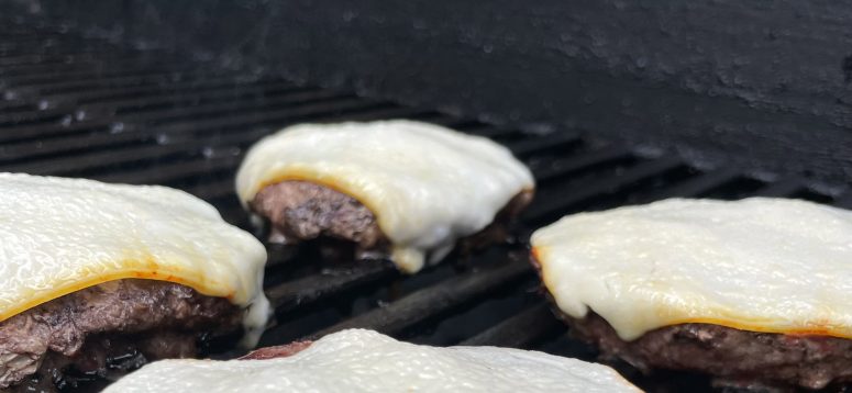 Four single patty burgers topped with cheese cooking on a grated grill.
