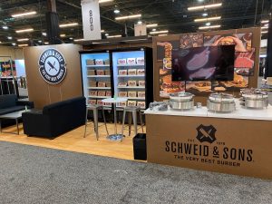 Tradeshow booth showing sample stations, a high-top table and behind it a cooler with products being displayed.