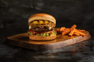 Burger with toppings including pineapple on a wooden board with a dark gray background. Sweet Potato Fries as a side.