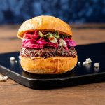 Burger on a big bun topped with bright red cabbage on a black plate.