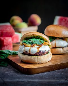 Burger with white mozzarella, peaches and basil on top drizzled with balsamic glaze on a brown cutting board.