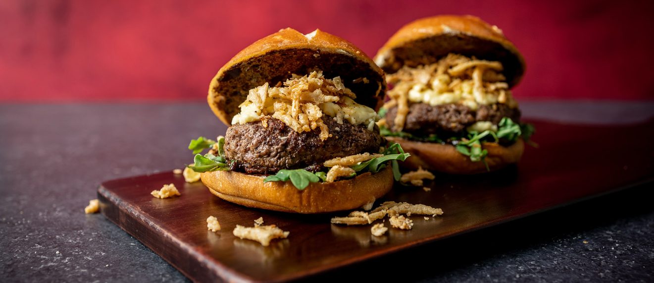 Two Burgers on a cutting board each topped with fried onion strings and mayo. Black Garlic Burger recipe