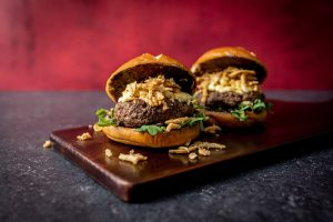 Two Burgers on a cutting board each topped with fried onion strings and mayo.