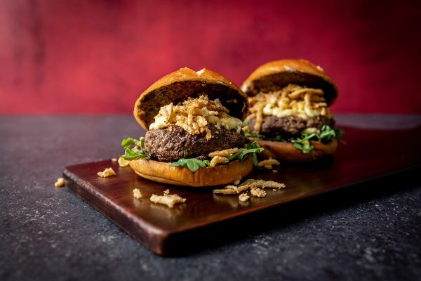 Two Burgers on a cutting board each topped with fried onion strings and mayo. Black Garlic Burger recipe