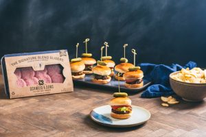 Cook up some Sliders for the ultimate at home tailgating party! Pictured are packaged Schweid & Sons Signature Blend Sliders with cooked sliders topped with tomatoes and pickles