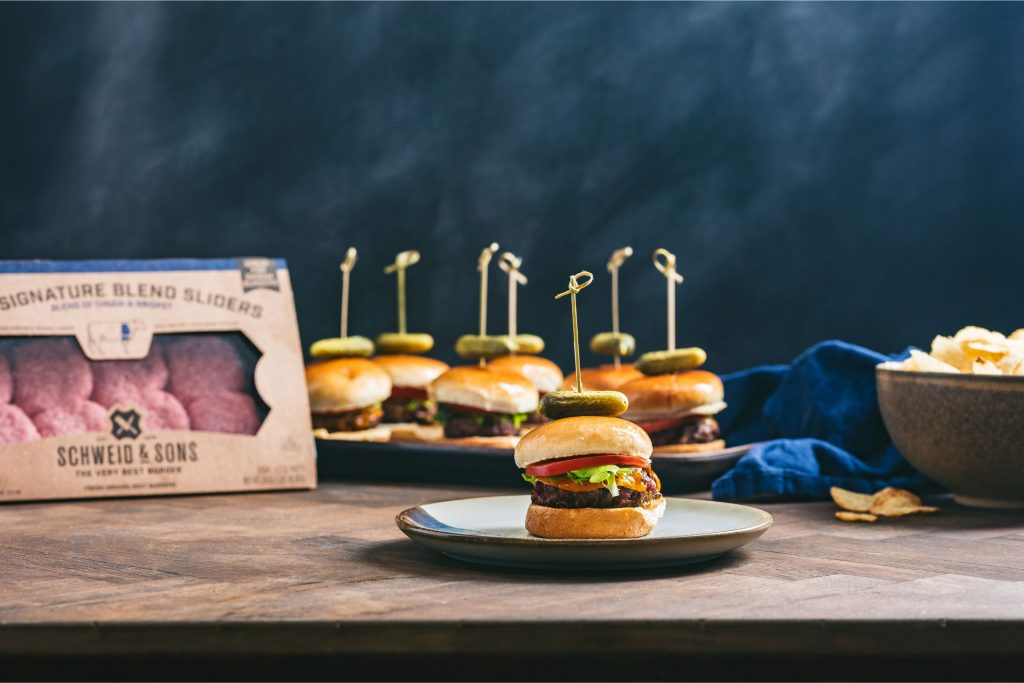 Cook up some Sliders for the ultimate at home tailgating party! Pictured are packaged Schweid & Sons Signature Blend Sliders with cooked sliders topped with tomatoes and pickles
