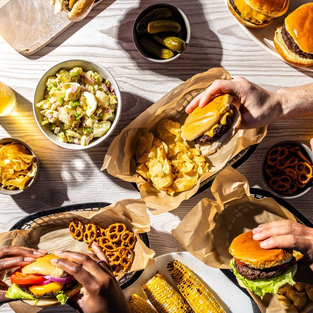 Schweid & Sons Burgers topped with a variety of tomatoes, onions, lettuce, and cheese with sides of salas, chips, pickles, corn, and pretzels