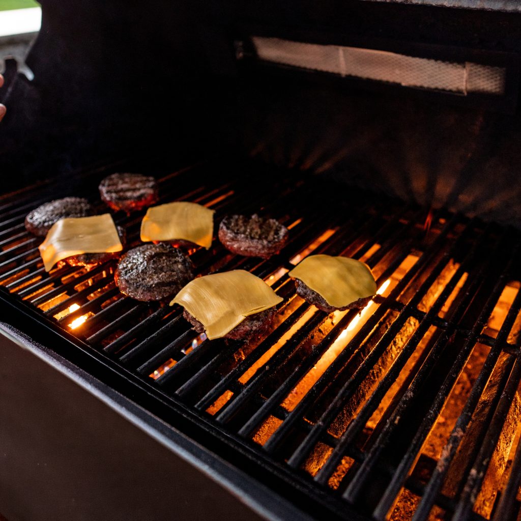 Schweid & Sons Burgers cooking on a grill topped with cheese
