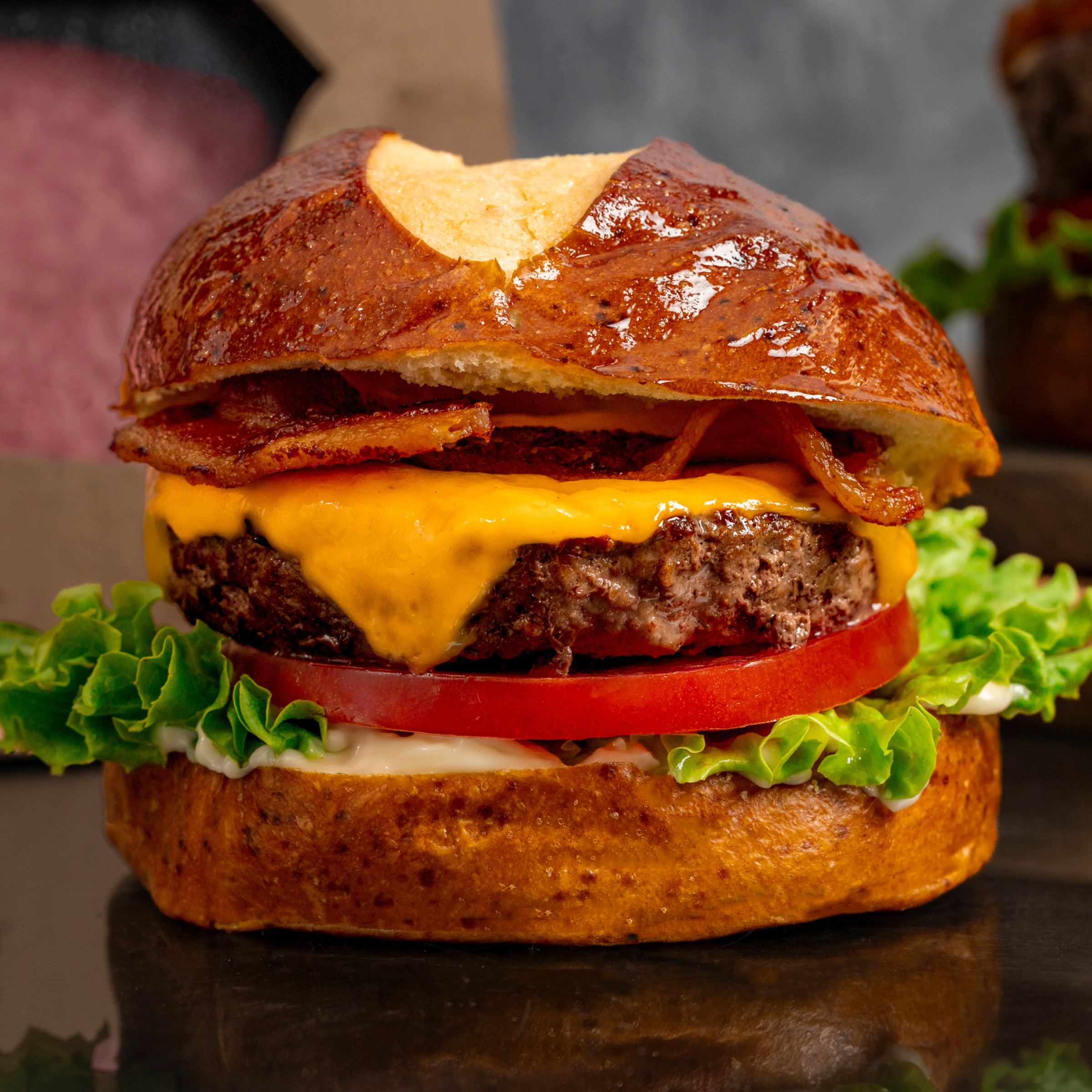 schweid and sons pretzel bun bacon cheeseburger topped with lettuce, bacon, cheddar cheese, and tomato all on a pretzel bun