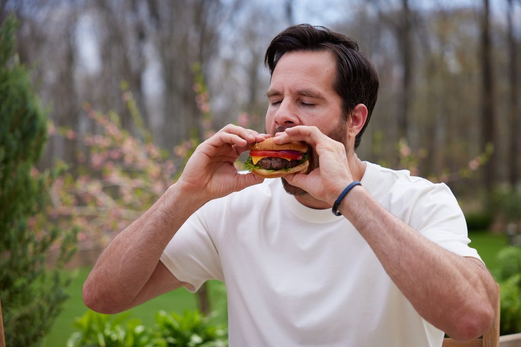Man biting into a schweid and sons cheeseburger topped with lettuce, tomato, onion and lettuce